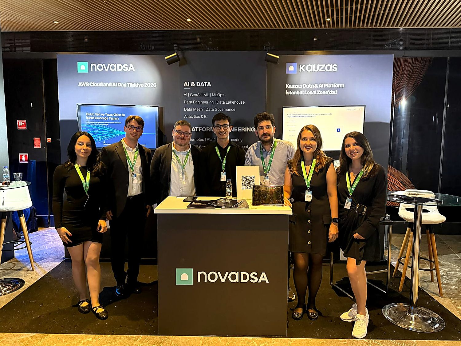 Novadsa ekibi AWS Cloud & AI Day 2026 standının önünde. Arkadaki panolarda Novadsa ve Kauzas logoları ile platformun kapsadığı başlıklar yazıyor.